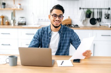 Confident clever positive arabian or indian stylish man with glasses, freelancer or product manager, sit at a desk at home in the kitchen, working on a project uses laptop, studying documents, smiling