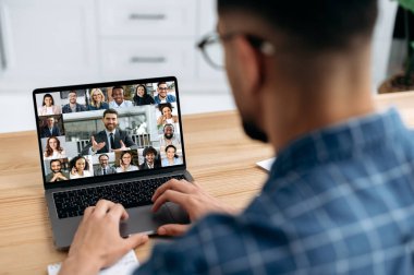 Over shoulder view at a laptop screen with different people, employees, business partners, male conducts online briefing, brainstorming, group conversation. Online video communication concept