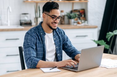 Positive confident arabian or indian young man with glasses, in stylish casual clothes, student, freelancer, company employee working remotely, sitting in the kitchen with a laptop, working on project