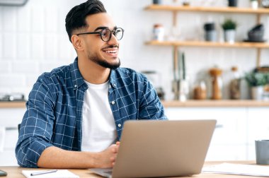 Handsome positive stylish indian or arabian young man with glasses, programmer, freelancer, working remotely, sitting at home in the kitchen with laptop, looking to the side, dreaming, smile happily