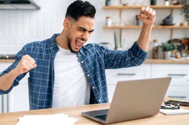Emotional amazed happy indian or arabian young man, trader, student, freelancer, sitting at table with laptop in the kitchen, enjoying big profit, victory, good news, gesturing with fists, smiling