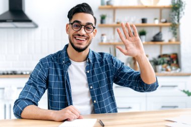Confident positive indian or arabian man, sit at desk at home, talk on webcam, having video call or conversation with client or colleague, mentor record training course, waves hand, smiling friendly