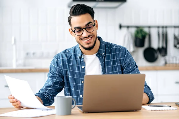 Work from home. Smart arabian or indian stylish man, freelancer, programmer, manager, working remotely, sit at a desk at home in the kitchen, working on a project uses laptop, studying documents,smile