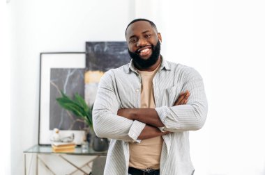 Positive confident handsome successful african american young man in stylish casual wear, with wireless headphones, standing indoors with crossed arms, looking at the camera, smiling friendly