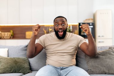 Excited joyful attractive young african american man, sitting on the sofa in living room, excited for success with arms raised, celebrating victory smiling happily, screaming. Winning concept