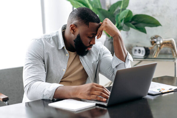 Sad exhausted african american young man, company employee, sitting at a desk, looks tired and stressed, experience headache, migraine, needs rest, closed his eyes. Overworked from work, deadline
