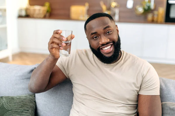 Happy African American guy, sit on sofa at home, holding a glass of clean water in hand, looks at camera, smiles. Handsome black guy leads healthy lifestyle, monitors the balance of water in the body
