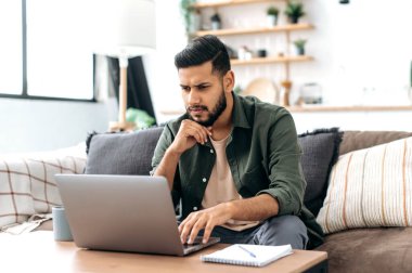 Focused smart arabian or indian young man in stylish casual clothes, freelancer, web designer, sitting in the living room on the sofa, working on a laptop on a project, looking at the screen intently