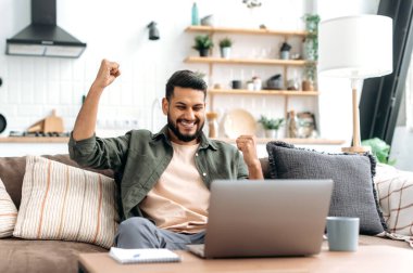 Amazed excited arabian or indian man, freelancer, sits on a sofa in living room, looks at laptop screen, gesticulating with hands, celebrates success, rejoice in big profit, received good news, smiles
