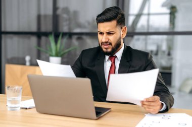 Disappointment stressed indian or arabian business man, sit at work desk with laptop in modern office, works with documents, upset with financial report, analyzes the result of work, feels frustrated