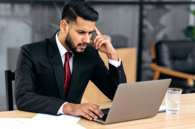 Confused puzzled indian or arabian young businessman, company ceo, in a suit, look at laptop screen in confusion, see unexpected message or news, feel stress, disappointment facial expression