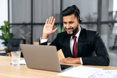 Successful confident indian or arabian businessman, company ceo, with headset, look at laptop screen, waves hand, talking with business client or partner online, explain business strategy and plan