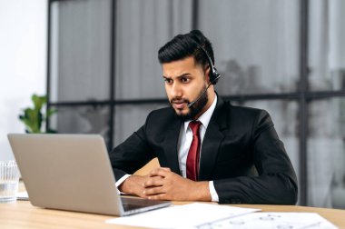 Puzzled stunned indian or arabian young businessman, company ceo, with headset, in a suit, look at laptop screen in confusion, see unexpected message or news, surprised emotional facial expression