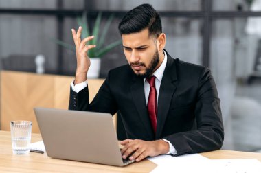 Disappointment facial expression. Confused puzzled indian or arabian young businessman, company ceo, in a suit, look at laptop screen in confusion, see unexpected message or news, feel stress