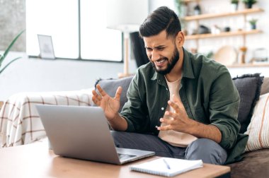 Stunned shocked indian or arabian guy, freelancer or student, sits on sofa in living room, looking disappointment at the laptop screen, stressed from the failure of a deal or project, in desperation