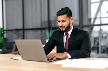 Focused intelligent confident arabian or indian successful businessman, company ceo, in formal suit, sit at work desk with laptop in modern creative office, concentrated looks at laptop screen