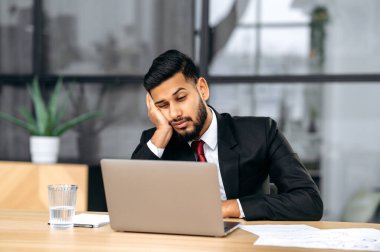 Fatigue, chronic sleep deprivation. Sleepy arabian or indian business man in a suit, sit at workplace in office with a laptop, put his head on his hand, fell asleep during work, feels tired,overworked