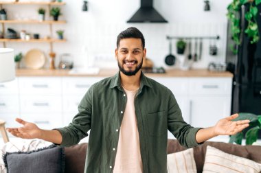 Happy handsome positive modern indian or arabian guy, in casual stylish clothes, stand at home in the living room on the background of the kitchen, spreads his arms to the side, looks at camera, smile