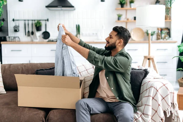 Excited arabian or indian man in casual clothes, sitting on a sofa in the living room, unpacking his parcel, taking out clothes from the cardboard box, looking at her happily, satisfied with new wear