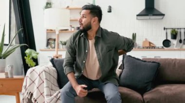 Problems with the back, spine, sciatica. Sad Arabian or Indian man, dressed in casual wear, trying to get up off the couch, experiencing acute pain in the back, lower back, has chronic joint problems