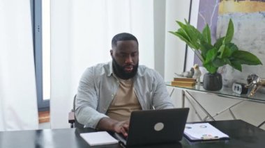 Amazed happy emotional african american stylish man, trader, student, freelancer, sitting at table with laptop in the office, enjoying big profit, victory, good news, gesturing with fists, smiling