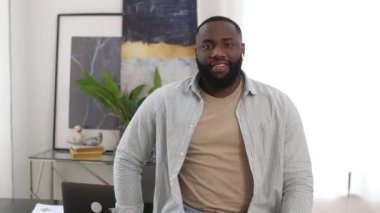 Positive confident handsome successful african american young man in stylish casual wear, with wireless headphones, standing indoors with crossed arms, looking at the camera, smiling friendly