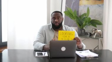 Sad unhappy unemployed african american man, sit at a desk in office, lost his job, holds a sign with the inscription looking for a job, hopes to get a new job, asks for support, sadly looks at camera