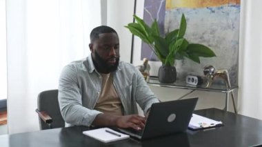 Sad exhausted african american young man, company employee, sitting at a desk, looks tired and stressed, experience headache, migraine, needs rest, feel stressed. Overworked from work, deadline