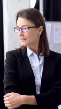 Vertical video portrait of pretty, elegant business woman in formal clothes, with glasses. Successful caucasian brunette woman, stands in the office with arms crossed, looks at camera, smiles friendly