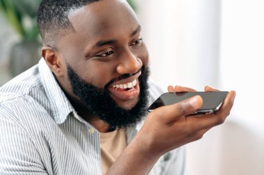 Recording a voice message. Wireless technology. Close up of a positive african american man holding his smartphone near his face, talking on speakerphone, recording audio message, smiling