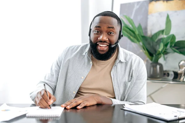 Confident african american man with headphones, business leader, company employee, having online business conversation, listens to the interlocutor, conducts consultation, takes notes, smiles