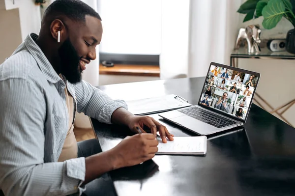 Group video meeting with multiracial people. Friendly african american man, sit at a desk in, having video meeting with colleagues, conducts financial brainstorm with business partners, takes notes