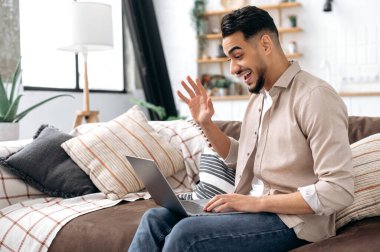 Cheerful positive arabian or indian young stylish modern man, spending time at home, sitting on sofa in living room, talking on video call with friends, family or colleague, waves hand, smiling