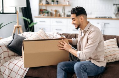 Excited mixed race young man opening big carton box, unpacking internet store order at home, happy with fast delivery service and the goods which received, smiling, happy facial emotions