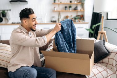 Excited happy indian or arabian guy opening big carton box, unpacking internet store order at home, contented with fast delivery service and the goods which received, smiling and feel satisfied