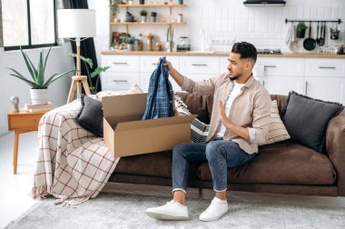 Disappointment arabian or indian stylish man, sitting on the sofa in the living room, unpacking his parcel, taking out clothes from the cardboard box, looking at her in frustration, dissatisfied