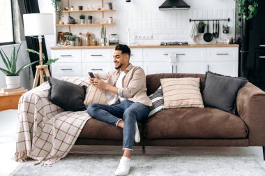 Online messaging, wireless technology. Happy indian or arabian guy, sit in modern living room on comfortable sofa, using his smartphone, browsing and messaging in a social media, surfing web, smiling