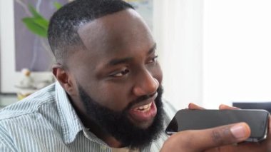 Recording a voice message. Wireless technology. Close up of a positive african american man holding his smartphone near his face, talking on speakerphone, recording audio message, smiling