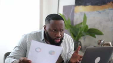 Confused puzzled african american business man, freelancer or company ceo, working with documents, sit at desk in modern office, working on a project uses laptop, studying financial graphs, stunned