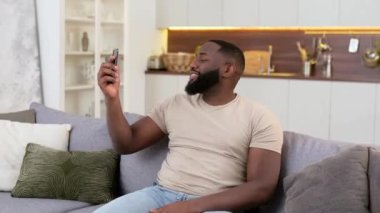 Joyful african american young stylish man sitting on sofa in living room, using his smart phone, talking on video call, having online conversation with friends in social networks, smiling