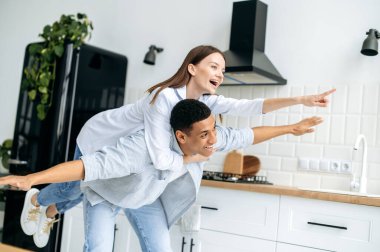Cheerful happy couple in love, caucasian girl and hispanic guy, dressed in a modern casual clothes, have fun together at home in the kitchen, laugh, enjoy the weekend, experiencing positive emotions