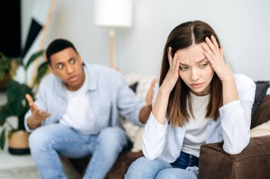 Misunderstanding in relationships, jealousy, quarrel. Mixed race couple sitting on a sofa in the living room, an angry man yells at a woman, feels angry, frustrated sad girl turned away from him