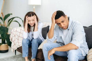 Quarrel, resentment against each other. Annoyed sad spouses, a multiracial couple, are sitting on a sofa in the living room, offended by each other after a showdown and shouting