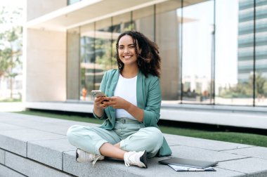 Joyful beautiful successful young mixed race woman, wearing stylish clothes, sitting outdoors, using her smartphone, chatting on social networks, browsing websites, looking at the camera, smiling
