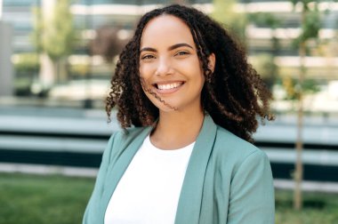 Portrait of beautiful positive confident successful hispanic or brazilian young woman with curly hair, in stylish elegant clothes, standing outdoors, looking at the camera smiling sincere smile