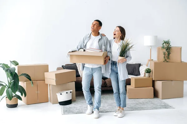 Enthusiastic joyful young couple in love, a caucasian woman and a mixed race man, are standing in their new home, happily looking around their new accommodation. Concept of moving, buying real estate