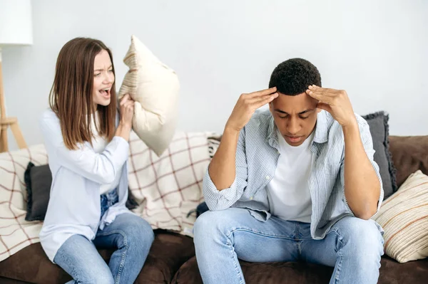 A married multiracial couple sorts things out while sitting in the living room on a sofa, a caucasian woman screams at her hispanic husband. Misunderstanding in relationships, jealousy, quarrel