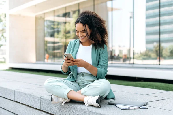 Online messaging. Happy lovely young mixed race woman, wearing stylish clothes, sitting outdoors, using her cellphone, chatting on social networks, browsing internet, answers email, smiling