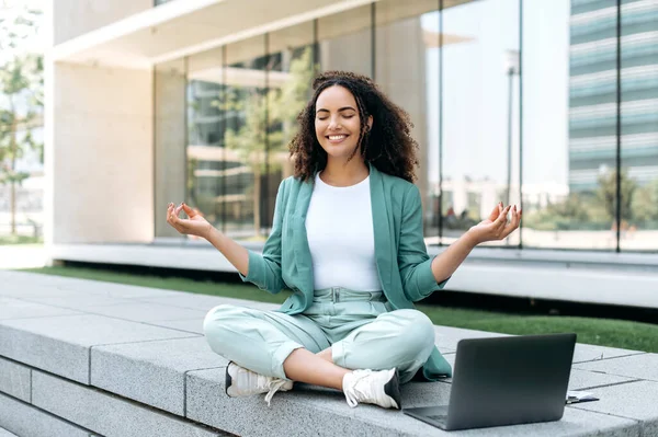 Break during work. Calm relaxed curly haired hispanic or brazilian young woman, office worker, sits in a lotus position outdoors near the business center, meditates, relaxes, closed her eyes, smile