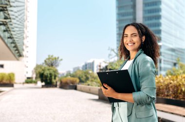 Successful positive confident young brazilian or hispanic curly haired business woman, real estate agent, manager, holds documents, stand outdoors near business center, looks at camera, smile friendly